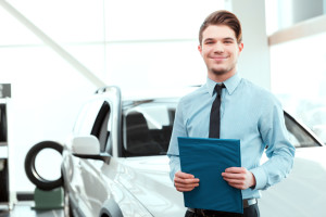 Perfect car dealer. Portrait of handsome young car sales man in formalwear holding a clipboard and smiling at camera in a car dealership